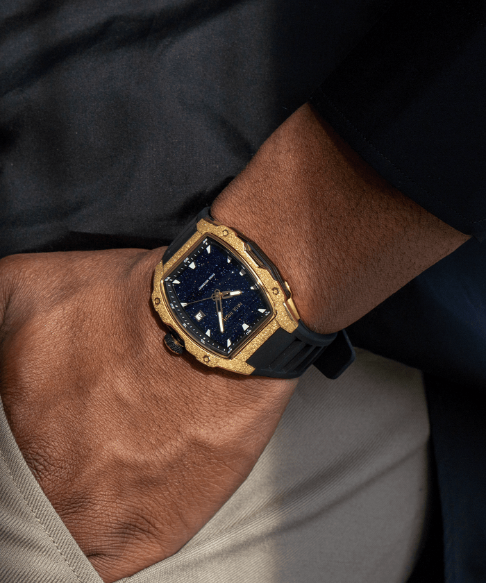 Tonneau-shaped Paul Rich watch with blue aventurine dial and gold frosted bezel worn on wrist, paired with beige pants and black top.