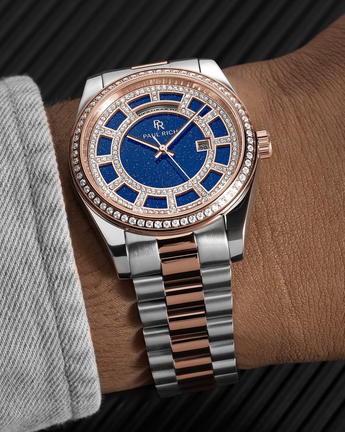 Rose gold and silver two-tone watch with a blue aventurine dial, pavé bezel, and day–date display shown on a wrist with a brushed metal bracelet.
