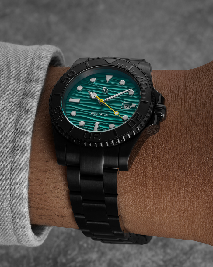 Black stainless steel watch with a teal wave-textured dial shown on a wrist, highlighting the bracelet, bezel, luminous markers, and yellow seconds hand.