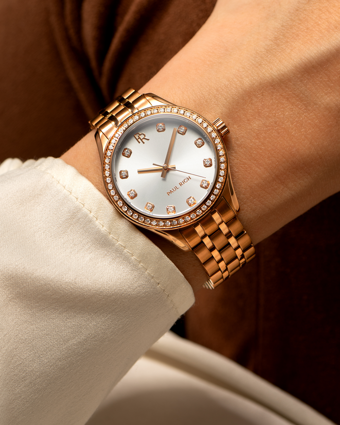 Close-up of a rose gold Paul Rich watch with diamond hour markers and a gem-set bezel, worn on a wrist with a cream-colored sleeve.