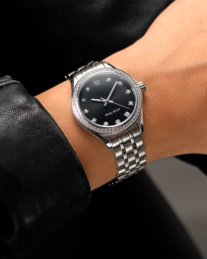 Silver-tone Paul Rich watch with black sunray dial, diamond hour markers, and gem-set bezel, worn on wrist with a black leather sleeve.
