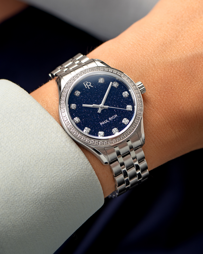 Silver-tone Paul Rich watch with a glittering deep blue aventurine dial, diamond hour markers, and gem-set bezel, worn on wrist with a light blue cuff.