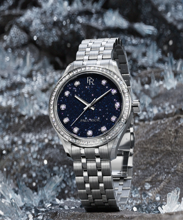 Silver-tone Paul Rich watch with deep blue aventurine dial, diamond hour markers, and gem-set bezel, displayed upright on sparkling crystal rocks.