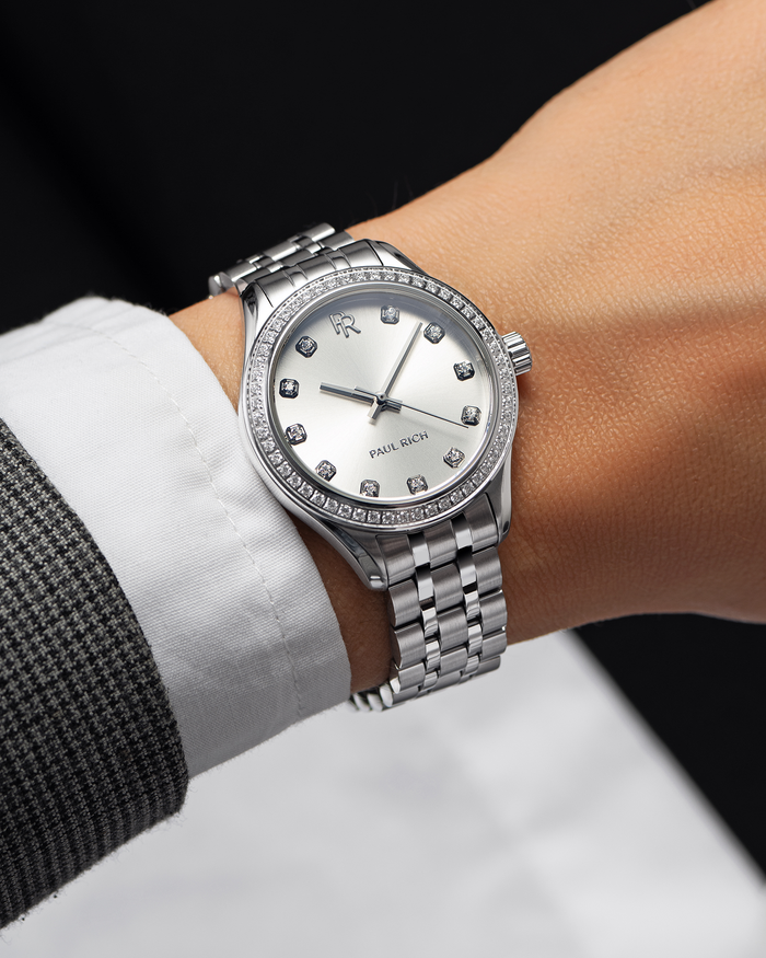 Silver-tone Paul Rich watch with a silver sunray dial, diamond hour markers, and gem-set bezel, worn on wrist with a white shirt and grey check blazer.