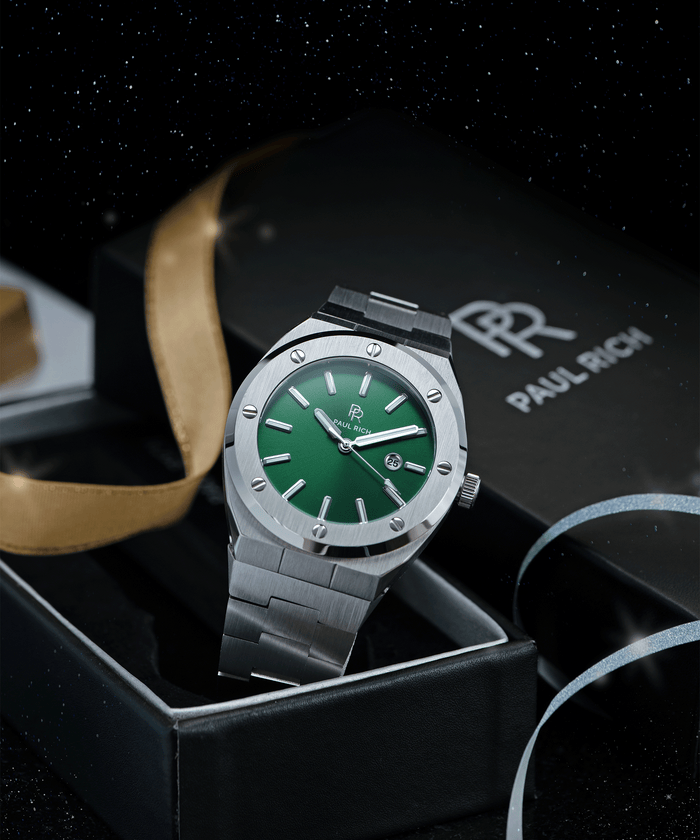 Paul Rich brushed silver watch with green dial and date window, displayed in open gift box with gold ribbon and starry night background.