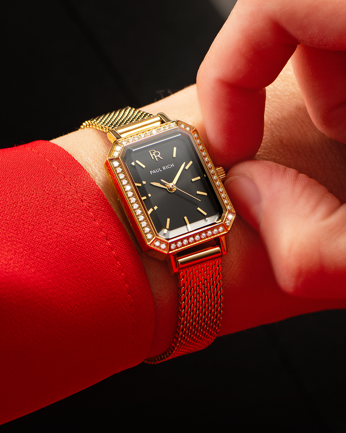 Gold-tone Paul Rich watch with black sunray dial, gem-set octagonal bezel, and gold mesh strap, worn on wrist with a red sleeve while a hand adjusts the crown.