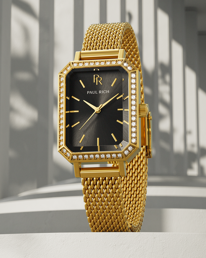Gold-tone Paul Rich watch with black sunray dial, gem-set octagonal bezel, and gold mesh strap, displayed upright on a white surface with vertical shadow patterns in the background.