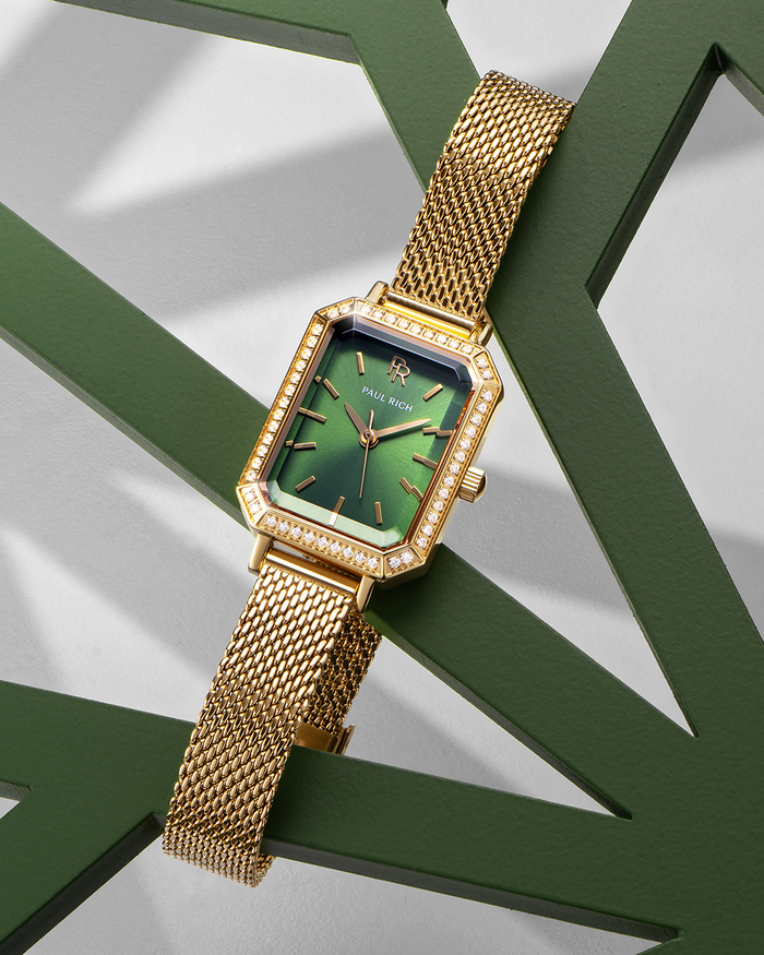 Gold-tone Paul Rich watch with a solid emerald green sunray dial, gem-set octagonal bezel, and gold mesh strap, displayed draped over a dark green geometric sculpture on a light background.