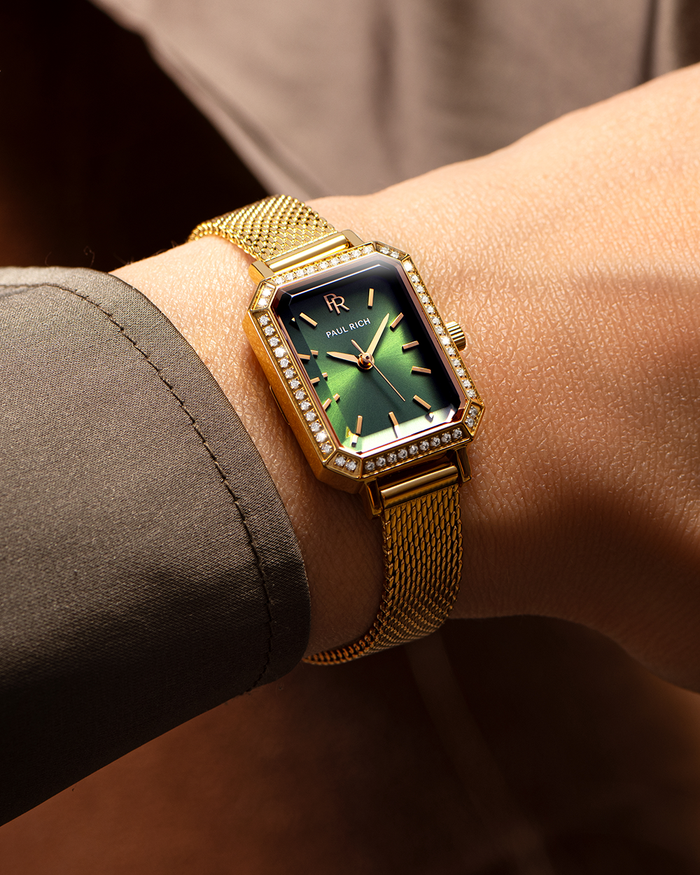 Gold-tone Paul Rich watch with emerald green sunray dial, gem-set octagonal bezel, and gold mesh strap, worn on wrist with a taupe-colored sleeve.