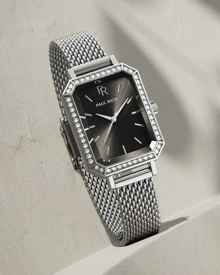 Silver-tone Paul Rich watch with black sunray dial, gem-set octagonal bezel, and silver mesh strap, displayed leaning against a textured beige wall with soft shadowing.
