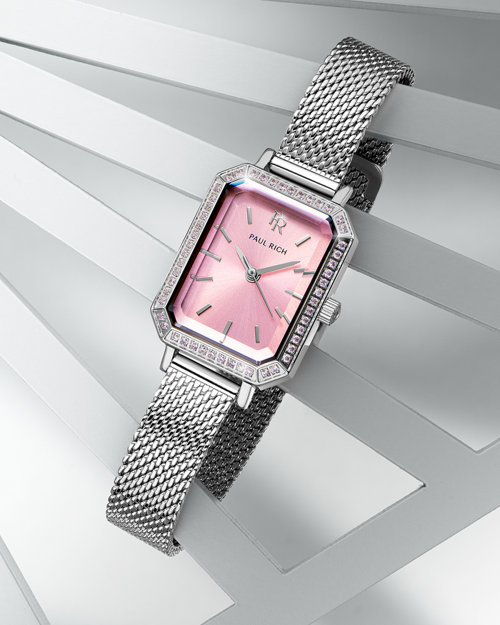Silver-tone Paul Rich watch with pink sunray dial, pink gem-set octagonal bezel, and silver mesh strap, displayed diagonally across layered white geometric panels.
