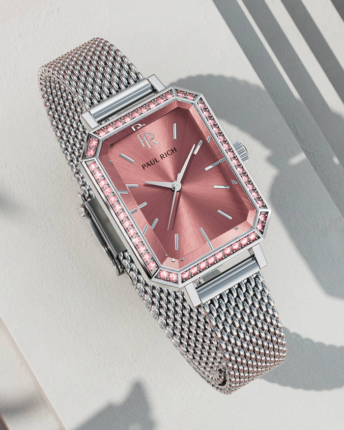 Silver-tone Paul Rich watch with rose pink sunray dial, pink gem-set octagonal bezel, and silver mesh strap, displayed on a textured off-white surface with sharp shadows.