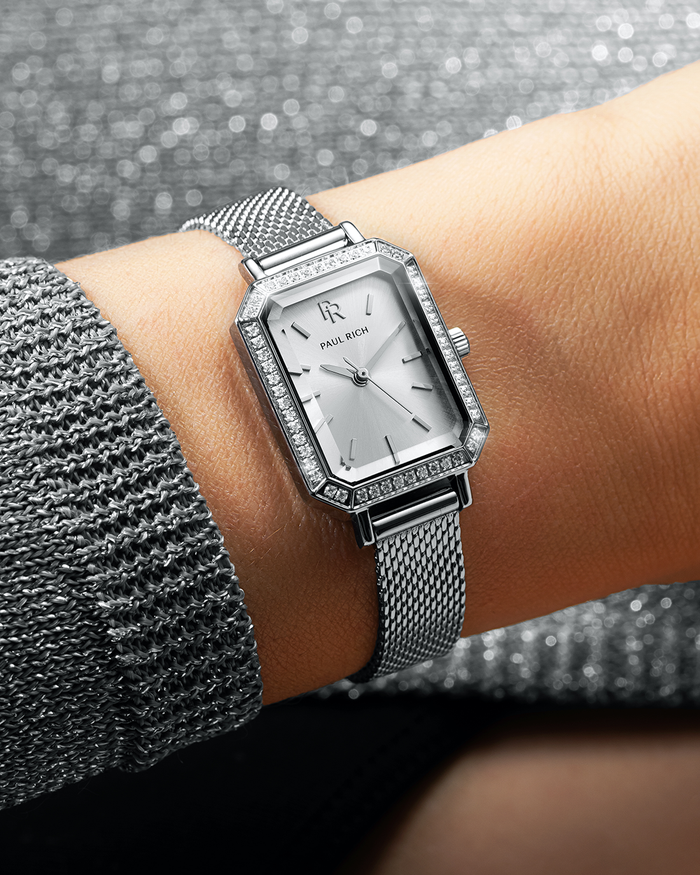 Silver-tone Paul Rich watch with silver sunray dial, gem-set octagonal bezel, and silver mesh strap, worn on wrist with a sparkling silver knit sleeve.