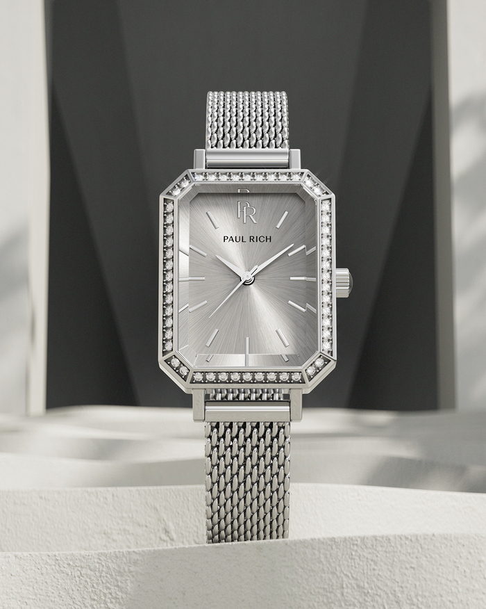 Silver-tone Paul Rich watch with silver sunray dial, gem-set octagonal bezel, and silver mesh strap, displayed upright on a textured off-white surface with geometric dark gray and white background.