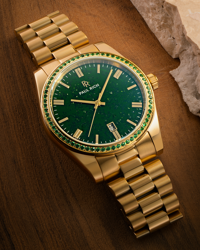 Gold watch with green aventurine dial and green gemstone-set bezel displayed on a wooden surface near a stone edge, showing the Paul Rich logo and date window.