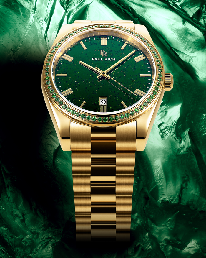 Gold watch with green aventurine dial and green gemstone-set bezel on a gold bracelet, displayed against a textured green background.