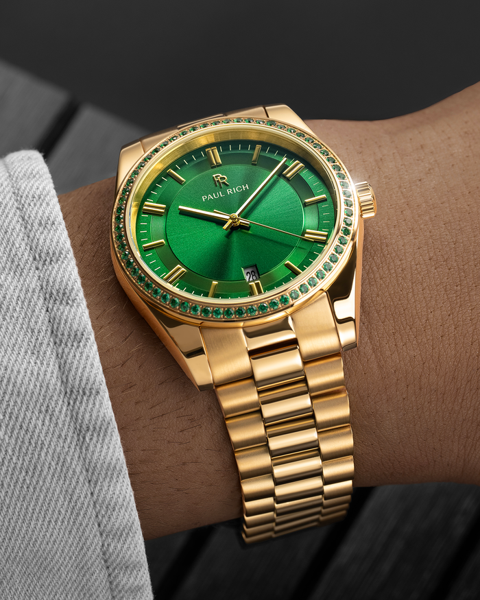 Gold watch with emerald-green dial and green stone bezel on a wrist wearing a light gray sleeve.