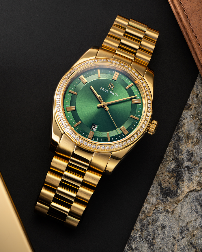 Gold-tone Paul Rich watch with green sunray dial, diamond-set bezel, and gold link bracelet, displayed on a flat lay surface with leather, stone, and matte black textures.
